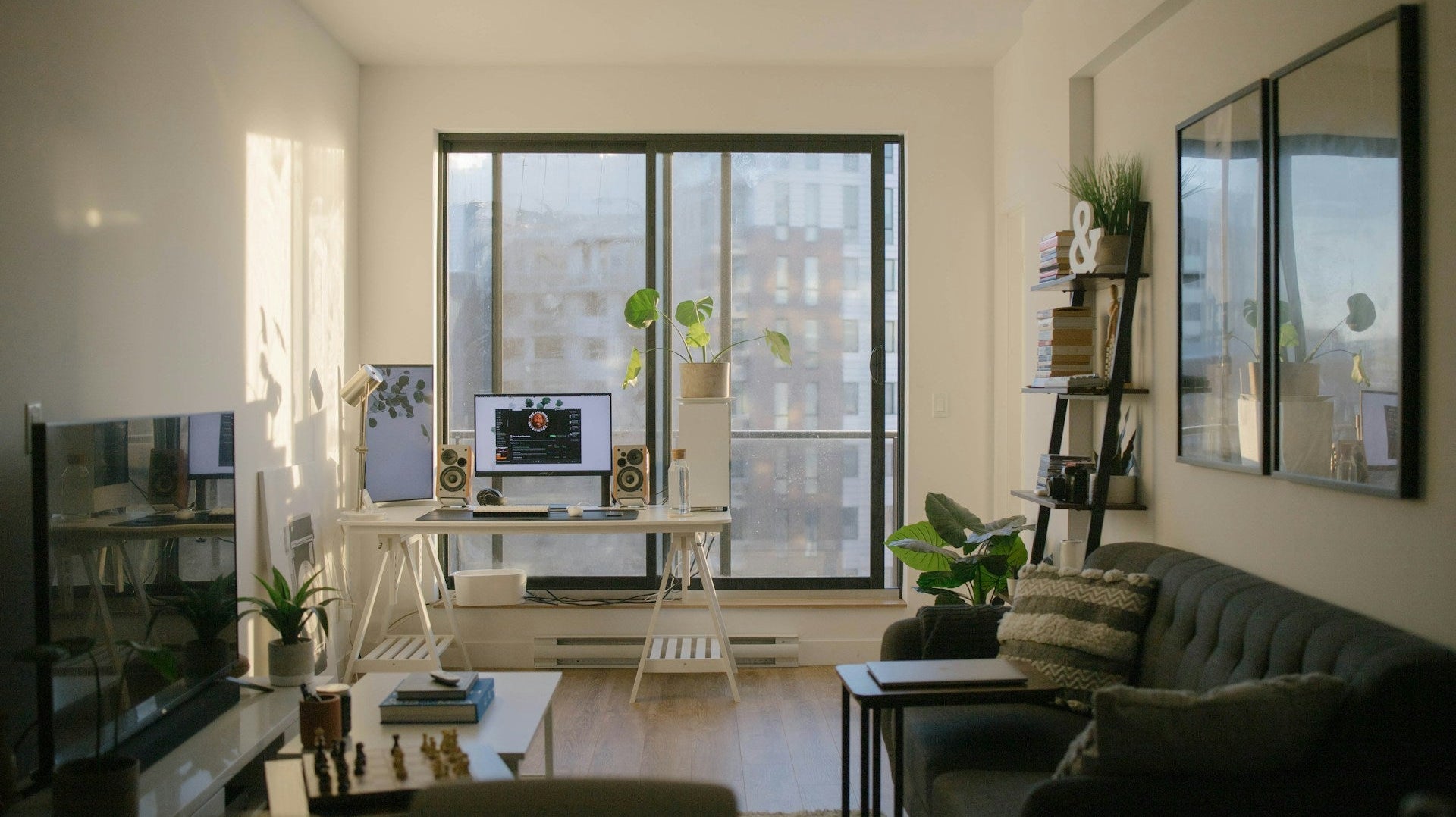 Stylish gaming setup living room with dual monitors, speakers, indoor plants, and cozy seating by a large window with city views.