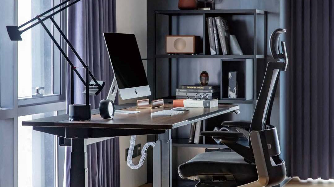 Modern home office setup featuring an ergonomic chair, standing desk, monitor, and organised cable management by a large window