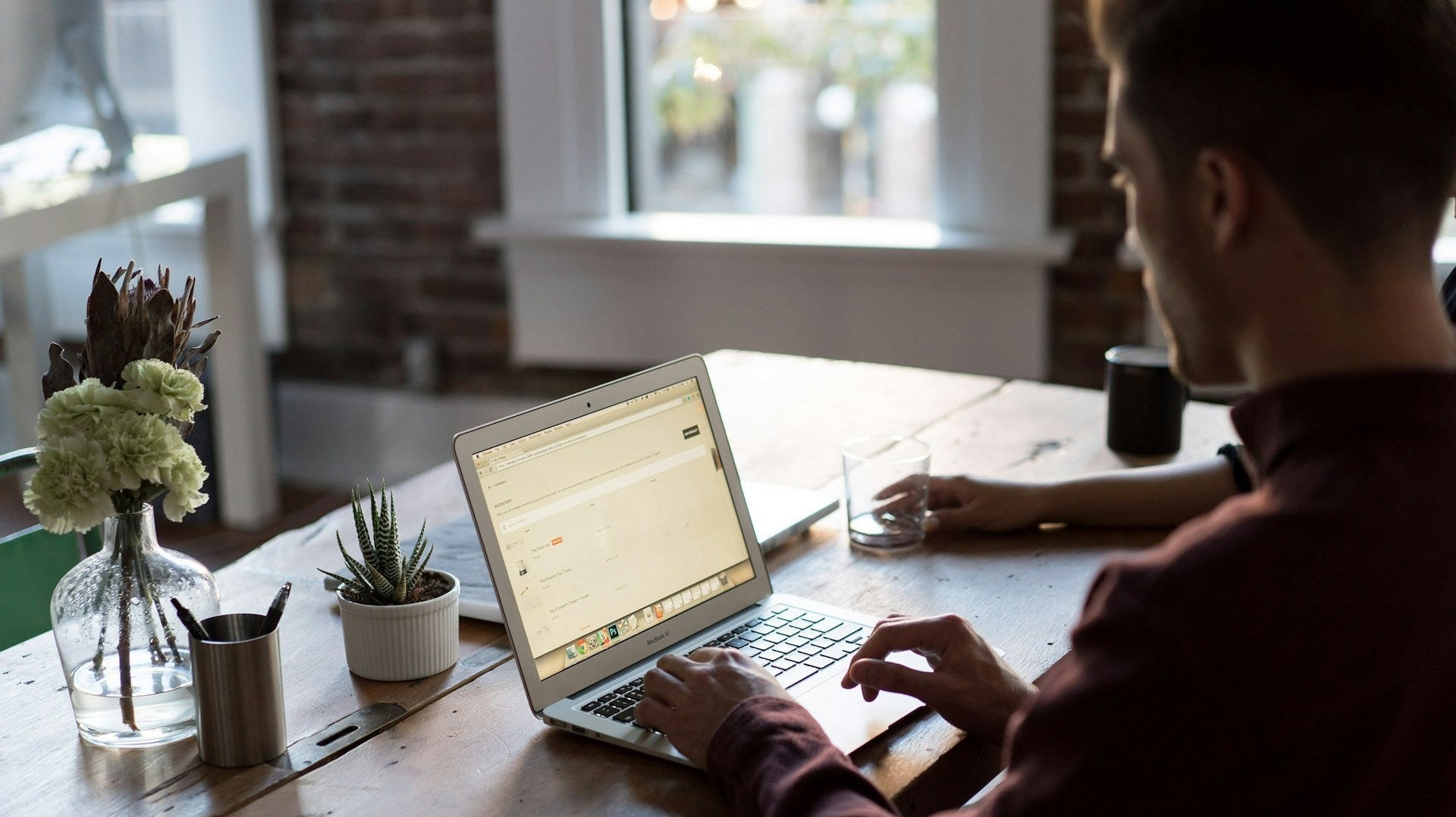 Man working on a laptop at a wooden desk with plants and natural light showcasing practical work from home setup ideas. Photo by Bench Accounting via Unsplash