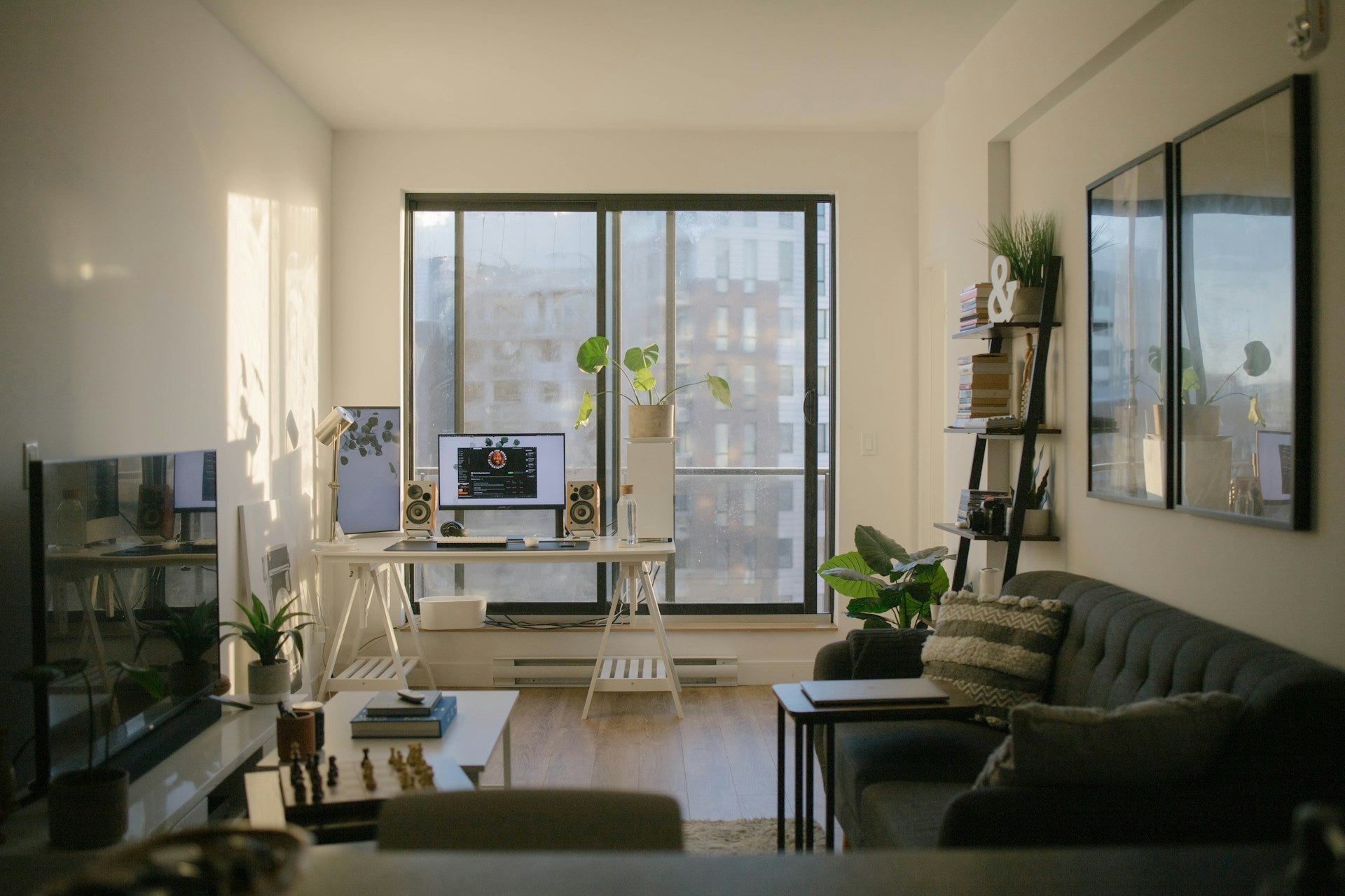 Stylish gaming setup living room with dual monitors, speakers, indoor plants, and cozy seating by a large window with city views.