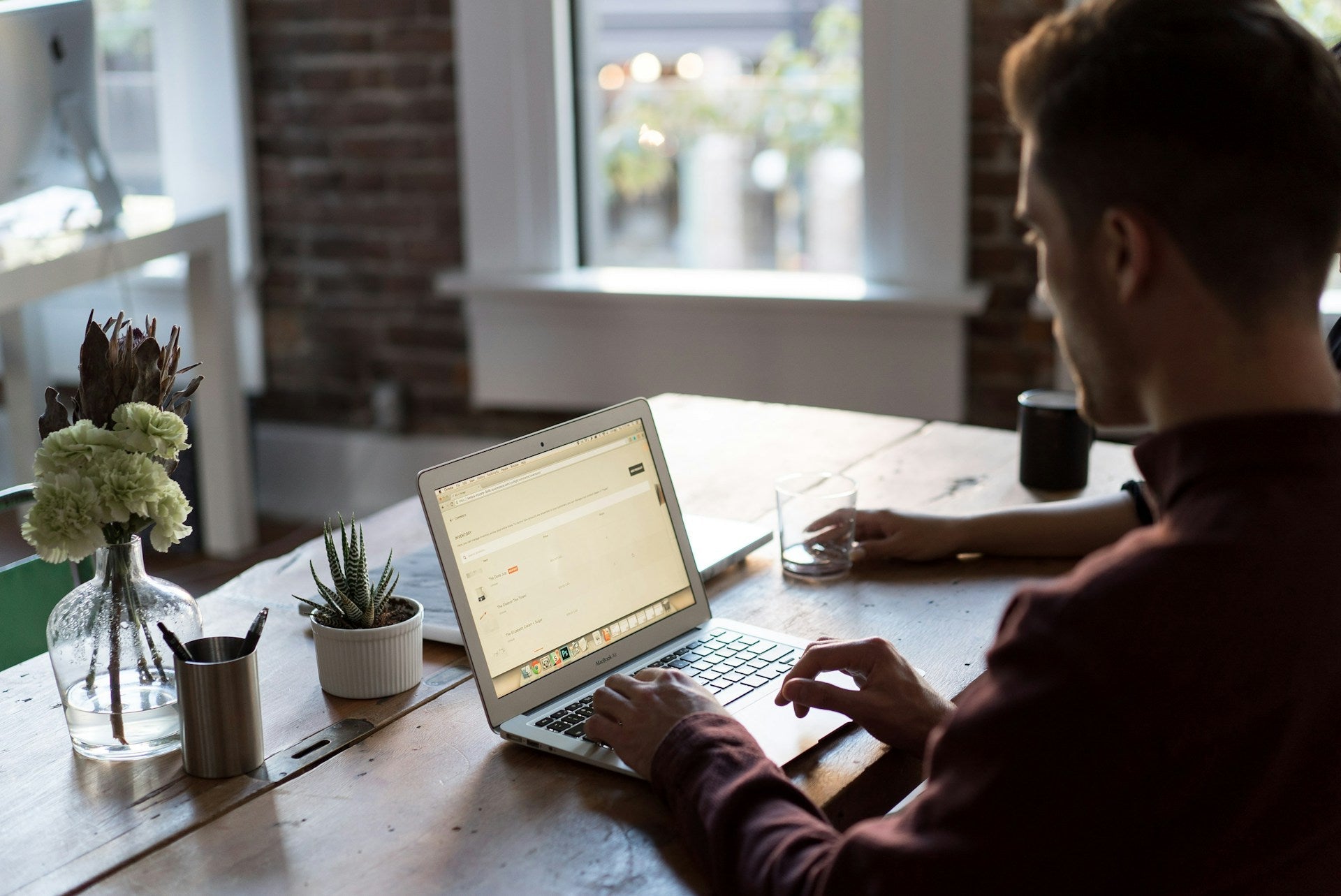 Man working on a laptop at a wooden desk with plants and natural light showcasing practical work from home setup ideas. Photo by Bench Accounting via Unsplash